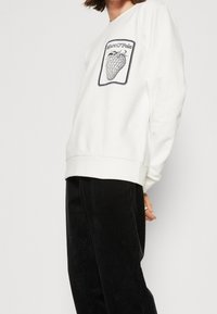 Vit sweatshirt med en jordgubbsdesign och svart kontur, med ribbade ärmslut och rund halsringning, tillsammans med svarta kordbyxor.