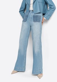 Jeans a gamba larga di un blu chiaro con vita alta, dotati di due tasche quadrate in tessuto denim e orli svasati. Tessuto liscio con una finitura sbiadita.
