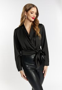 Sort satin indpakningstop med lange ærmer og detalje med bundet talje, parret med sorte tætsiddende bukser, der skaber en slank silhuet.