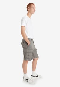 Pantaloni cargo grigi con multiple tasche, abbinati a una maglietta bianca e sneakers bianche con dettagli neri. Modello in profilo laterale.