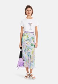 Weißes Baumwoll-T-Shirt mit einem Cocktail-Graphic, kombiniert mit einem bunten, gemusterten Midikleid in den Farben Grün, Lila und Pink, und einer lavendelfarbenen Tasche.