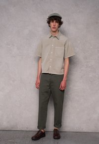 Korte mouwen, lichtbeige overhemd met knopen en een borstzak, gecombineerd met donkergroene tapered broek en bruine veterschoenen.
