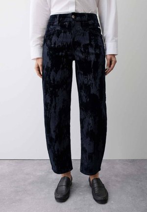Persoon die zwarte, hoog getailleerde fluwelen broek draagt, een wit overhemd met lange mouwen en zwarte instap loafers, staand op een grijze vloer.