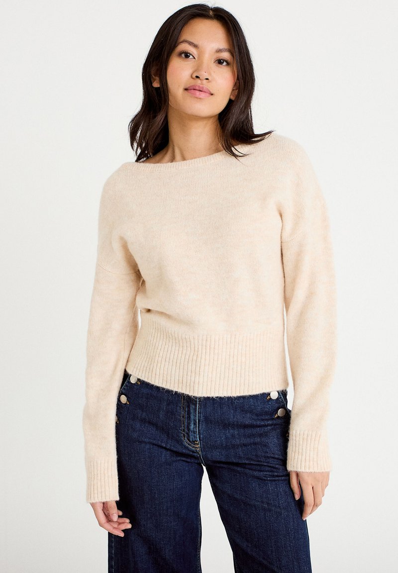 Frau mit dunklem Haar, die einen cremefarbenen Pullover und dunkelblaue Jeans trägt, stehend vor einem schlichten weißen Hintergrund.