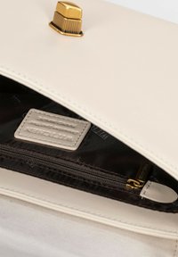 L'intérieur d'un sac à main en cuir crème présente un fermoir doré, une poche zippée et une étiquette de marque cousue sur un doublage sombre. Texture lisse.