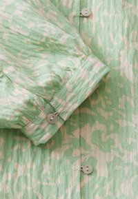 Manche de chemise à motifs verts et crème, avec poignet boutonné et boutons visibles à l'avant sur un tissu texturé.