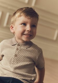 Polo beige con un patrón texturizado, tres botones y un cuello a juego. Lleva puesto un niño con el cabello corto y claro y una sonrisa.