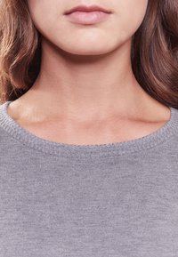 Zbliżenie dolnej części twarzy kobiety, szyi i obojczyka o jasnej skórze, noszącej szary dzianinowy sweter.