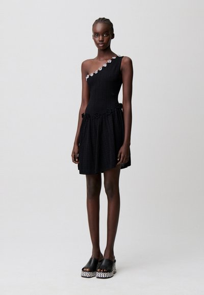 Donna che indossa un vestito nero senza maniche con bordo floreale su una spalla e in vita, abbinato a sandali platform neri con suola decorata.
