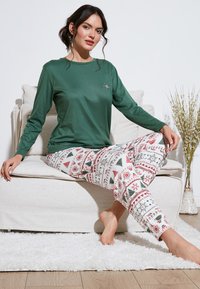 Cămașă verde cu mâneci lungi, cu un mic detaliu decorativ, asociată cu pantaloni de pijama albi, cu imprimeuri festive în nuanțe de roșu și verde, cu motive de copaci și fulgi de zăpadă.