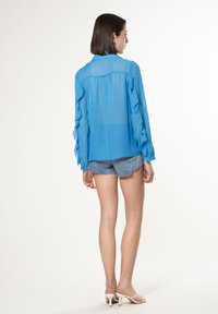 Doorzichtig blauwe blouse met gerimpelde mouwen, gecombineerd met denim shorts. De top heeft een kraag en een rechte snit, wat de lichte uitstraling benadrukt.