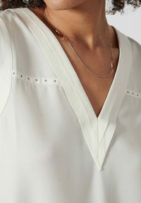 Blouse blanche avec un col en V, dotée de détails de couture verticaux et de petits pois décoratifs sur les épaules. Tissu lisse et léger.