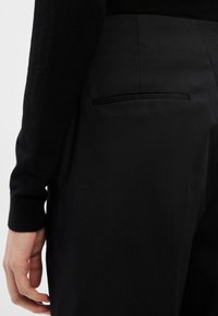 Κοντινό πλάνο ενός ατόμου που φορά ψηλόμεσα μαύρα παντελόνια με πίσω τσέπη τύπου welt και μαύρο μακρυμάνικο πουλόβερ, το χέρι χαλαρό δίπλα στο σώμα.