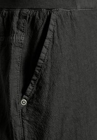 Gros plan d'une poche de pantalon en tissu noir texturé avec des coutures visibles et une petite rivet métallique dans le coin.