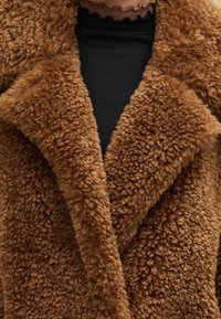 Abrigo de piel sintética marrón con una textura suave y esponjosa. Presenta un cuello con solapa y un frente superpuesto, sobre una camiseta negra.