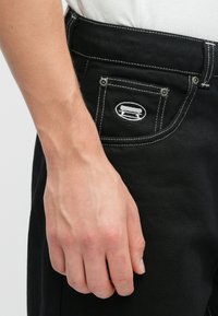 Černé džíny z denimu s bílým prošitím, s zadní kapsou s vyšitým logem. Záběr zblízka na ruku položenou na pase.