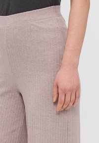 Pantalones de tela acanalada gris claro, con una cinturilla ancha y elástica. La vista en primer plano resalta la textura y el acabado suave. Mano colocada sobre la tela.