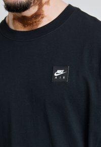 Black cotton T-shirt with round neckline, featuring a small white Nike Air logo patch on the left chest area. Soft, simple design.