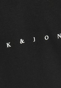 Tessuto nero con testo bianco "K & J" in un font pulito e minimale. La texture appare morbida e liscia.