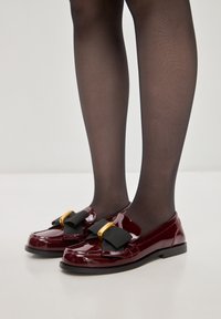 Mocassini in pelle verniciata bordeaux con un fiocco in tessuto nero e dettagli hardware dorati. Design a profilo basso con punta arrotondata e suola nera.