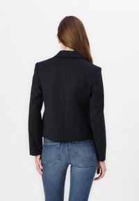 Czarna, krótka marynarka z kołnierzem o wyszczególnionym kształcie i długimi rękawami, wykonana z gładkiego materiału. Nosić na jasnoniebieskich dżinsach. Ukazany widok z tyłu.