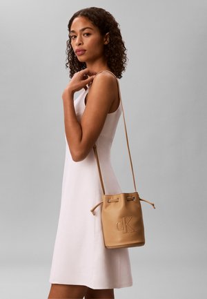 Young woman with curly hair wearing sleeveless white dress, carrying tan bucket bag with "cK" logo on shoulder against gray background.
