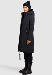 Sort polstret frakke med hætte, lynlåslukning, diagonalt quiltet design og en sort rund lomme. Båret med en sort hue og støvler.