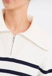 Sweater blanc zippé avec un col texturé et des rayures horizontales bleu marine. Il dispose d'une fermeture éclair argentée et de détails côtelés sur le col.