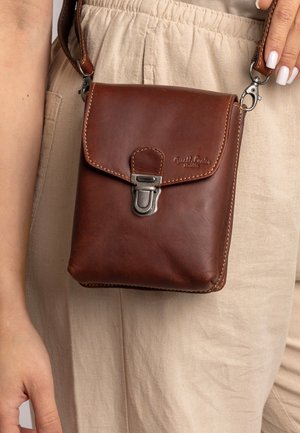 Borsa a tracolla in pelle marrone con chiusura a patta, hardware argentato, dettagli in cucitura e superficie testurizzata, indossata all'anca su un tessuto leggero.