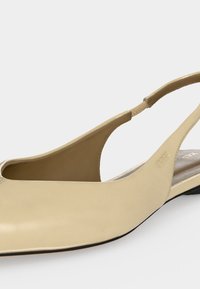 Chaussure plate beige en cuir à bride arrière avec bout pointu et bride élastique au talon sur fond blanc.