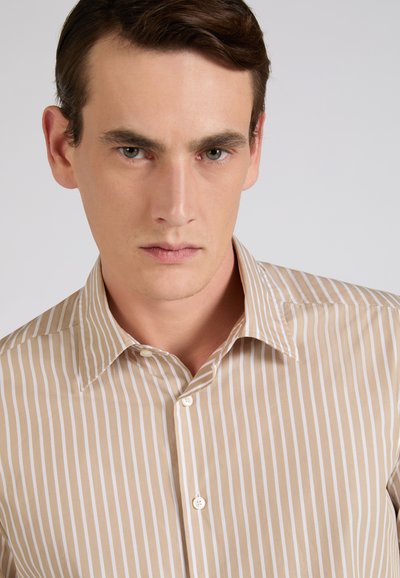 Jonge man met kort bruin haar draagt een beige-wit gestreept overhemd met verticale strepen tegen een effen grijze achtergrond.
