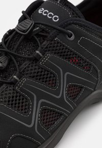 Chaussure de sport en maille noire avec une construction sans coutures, des accents rouges, un système de laçage ajustable et une semelle extérieure en caoutchouc avec une adhérence texturée.