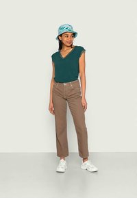 Haut teal à col en V avec des manches effilochées, pantalon droit beige, et un chapeau en seau à motifs bleus. Des baskets blanches complètent la tenue.