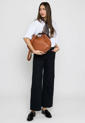 Young woman in white shirt and black pants holding a brown leather handbag, standing against a plain light background.