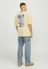 Lichtgele oversized t-shirt met een grafische print op de achterkant die een strandtafereld afbeeldt, gecombineerd met baggy blauwe denim cargo broeken.