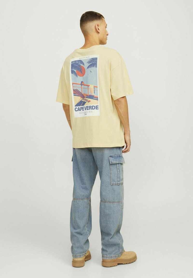 Lichtgele oversized t-shirt met een grafische print op de achterkant die een strandtafereld afbeeldt, gecombineerd met baggy blauwe denim cargo broeken.
