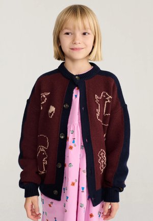 Jeune fille aux cheveux blonds coupés au carré, portant un cardigan bordeaux et bleu marine sur une robe rose à motifs floraux colorés, debout devant un fond uni.