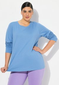 Světle modrý top s dlouhým rukávem a kulatým výstřihem. Má na rukávech nařasené detaily. V kombinaci s fialovými legínami. Hladká textura.