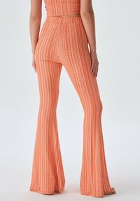 Uitlopende broek in oranje stof met verticale strepen en textuuraccenten. Heeft een hoge taille en rekbaar materiaal voor comfort.