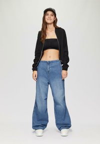 Zwarte zip-up bomberjack, crop top en losse blauwe denimspijkerbroek; witte sneakers; model staat op een neutrale achtergrond.