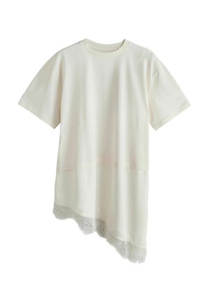 Kortærmet off-white lagdelt top med asymmetrisk blondekantet sømdetalje på en ensfarvet baggrund.