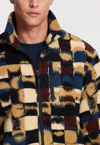 Giacca in pile con patchwork multicolore di beige, marrone, blu e nero, dotata di una tasca sul petto con zip e colletto alto.