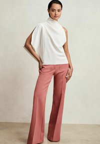 Top alb asimetric cu guler înalt și design drapat, asociat cu pantaloni roz evazați, croiți, cu buzunare la spate.