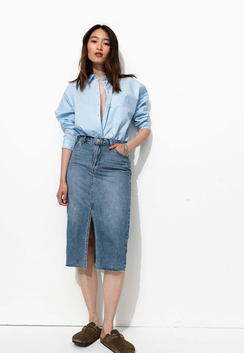 Chemise bleue claire à boutons, jupe en denim taille haute avec fente à l'avant, et sabots marron avec des accents cloutés sur fond blanc.