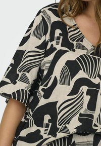 Haut à motifs noir et blanc avec un décolleté en V, des manches courtes et un mélange de formes et de lignes audacieuses sur un fond en tissu texturé.