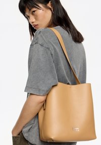 Borsa a spalla in pelle tan con una superficie liscia, caratterizzata da un design semplice e un logo impresso alla base. Manico sottile attaccato.