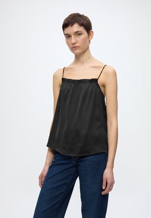 Γυναίκα με κοντά σκούρα μαλλιά που φοράει μαύρο σατέν τοπ χωρίς μανίκια με λεπτές τιράντες και μπλε τζιν παντελόνι, στέκεται μπροστά σε λευκό φόντο.