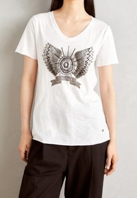 Vit bomullströja med v-ringning, prydd med en metallisk grafik av vingar och ett cirkulärt mönster, samt en liten logotyp vid fållen.