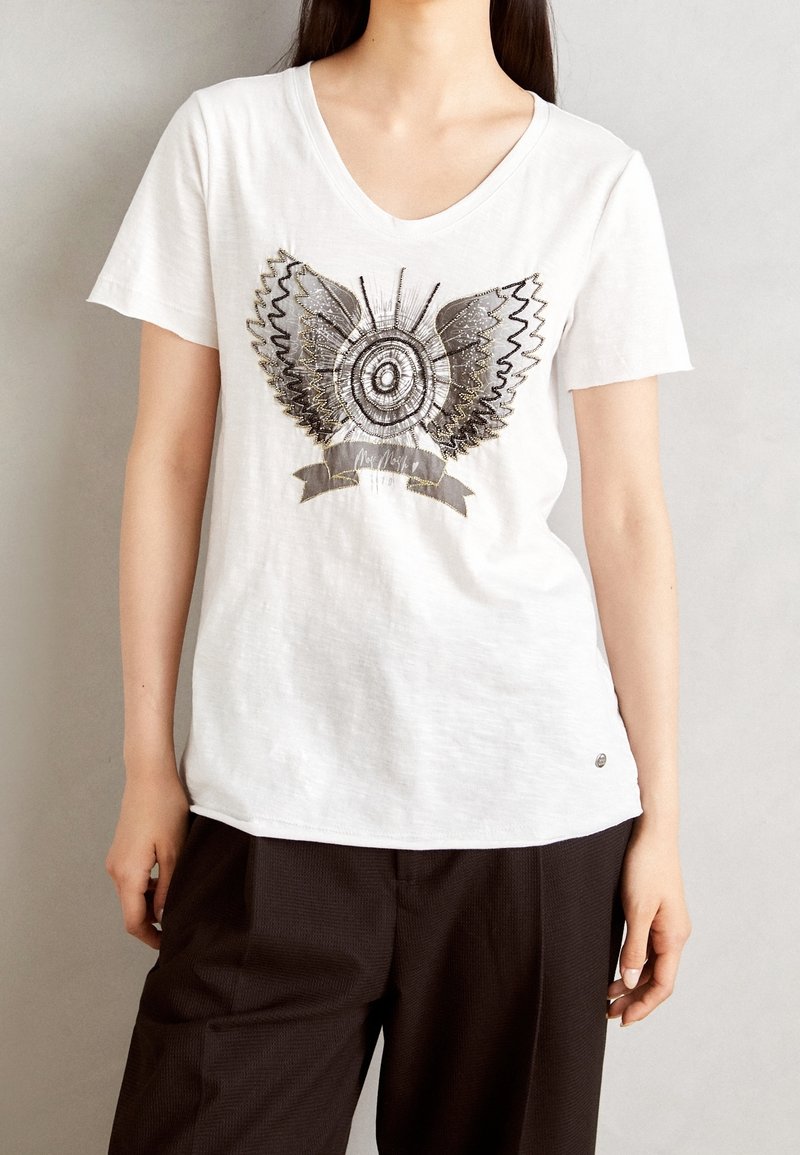 Vit bomullströja med v-ringning, prydd med en metallisk grafik av vingar och ett cirkulärt mönster, samt en liten logotyp vid fållen.