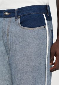 Gros plan sur un jean en denim bicolore bleu avec une poche supérieure foncée, une bande latérale blanche et une main visible reposant sur la cuisse.